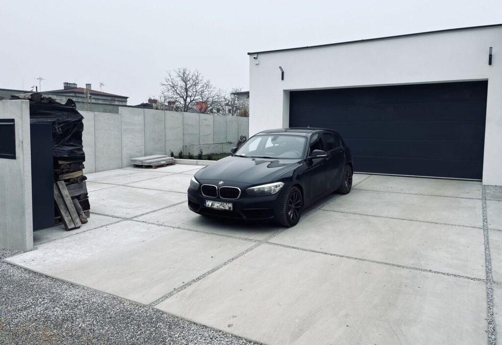 Czarny samochód na betonowym podjeździe. W tle garaż z czarną bramą wjazdową.